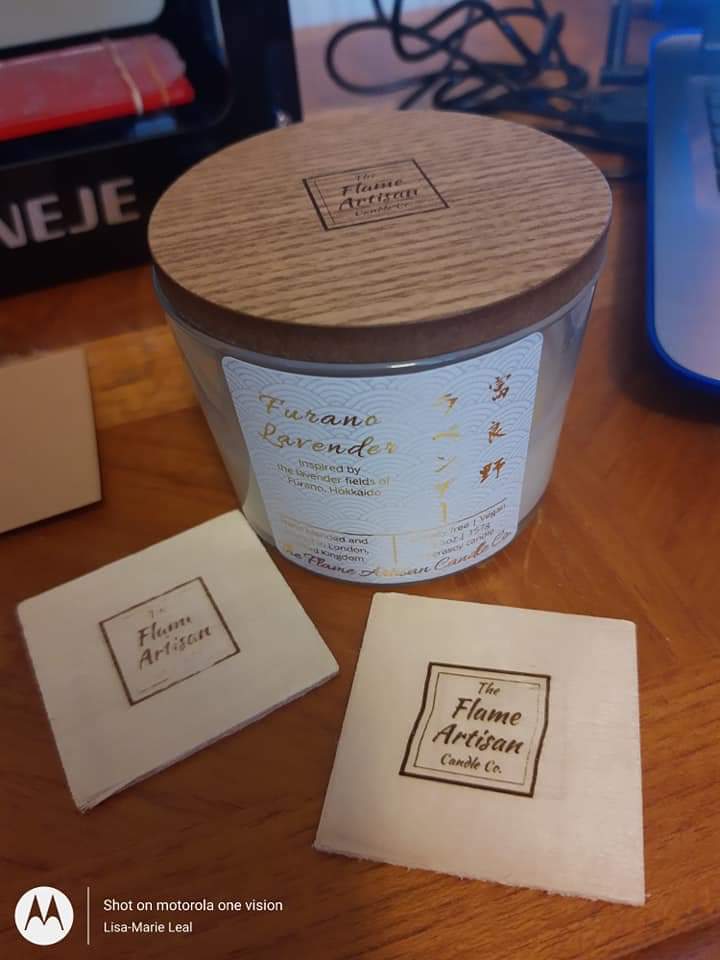 Candle with wooden lid and label on a wooden surface, with 'The Flame Artisan' branding.